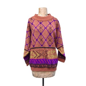 May include: A vintage sweater in shades of peach, purple, black, and gold, displayed on a mannequin. The sweater features a diamond pattern in purple, with black accents, and a geometric design at the bottom. The long-sleeved sweater has a ribbed collar.