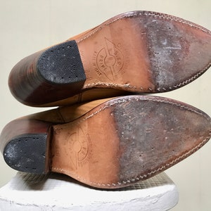 May include: A pair of tan leather cowboy boots, viewed from the soles. The boots have pointed toes, stacked heels, and rubber heel caps. The soles show wear and stitching. The boots are labeled "Made in Italy".