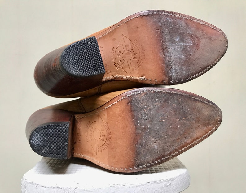 May include: A pair of tan leather cowboy boots, viewed from the soles. The boots have pointed toes, stacked heels, and rubber heel caps. The soles show wear and stitching. The boots are labeled "Made in Italy".
