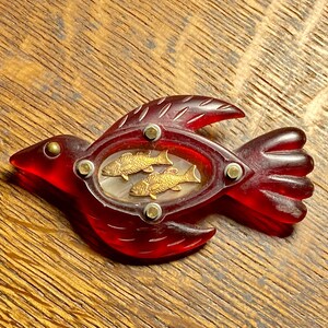 May include: A vintage red bird-shaped brooch with a clear, glossy finish. The brooch features a central oval design with two gold-colored fish. The bird's wings and tail are detailed, and the brooch is secured with small, round fasteners.