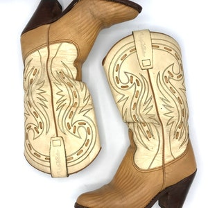 May include: A pair of beige and cream-colored cowboy boots. The boots have a pointed toe and a stacked heel. The shaft of the boots features decorative stitching and a pull strap. The heel is dark brown.