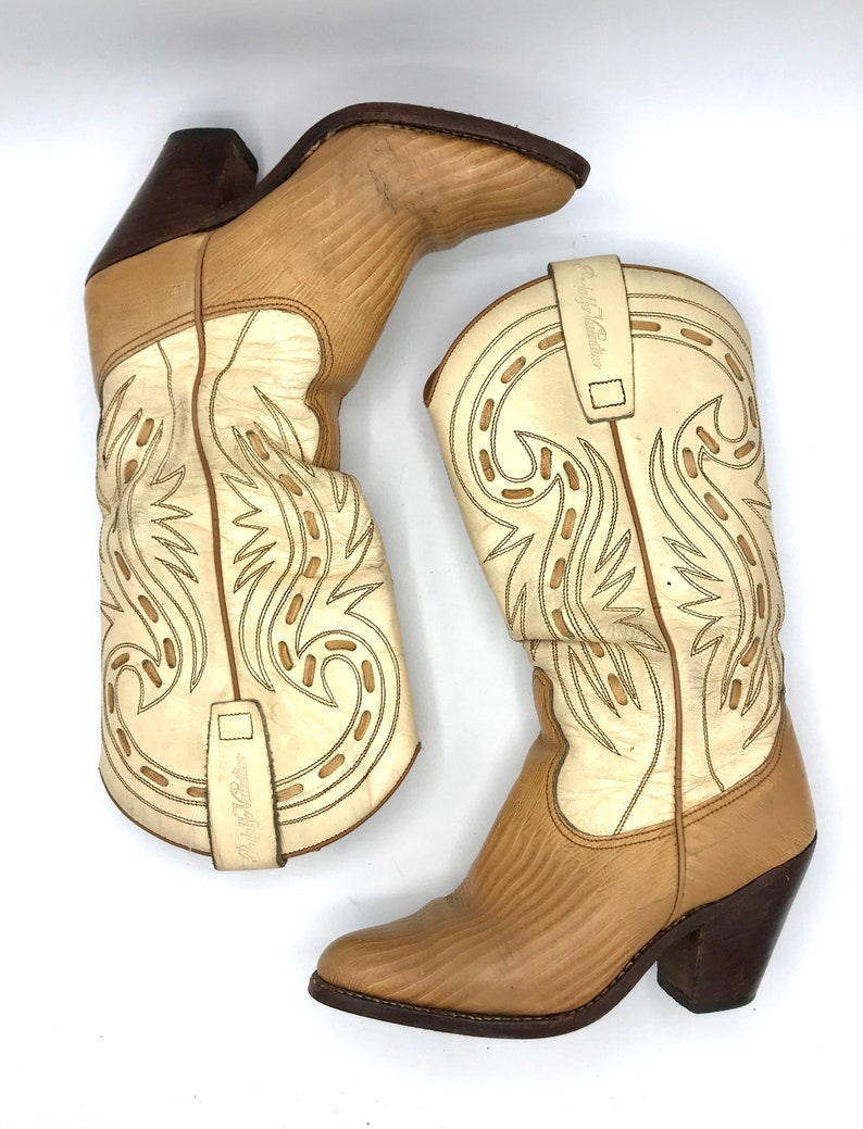 May include: A pair of beige and cream-colored cowboy boots. The boots have a pointed toe and a stacked heel. The shaft of the boots features decorative stitching and a pull strap. The heel is dark brown.