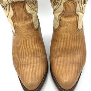 May include: A pair of cowboy boots with a light brown, textured leather design. The boots have a cream-colored upper with decorative stitching and a dark brown sole. The toe of the boot is pointed.