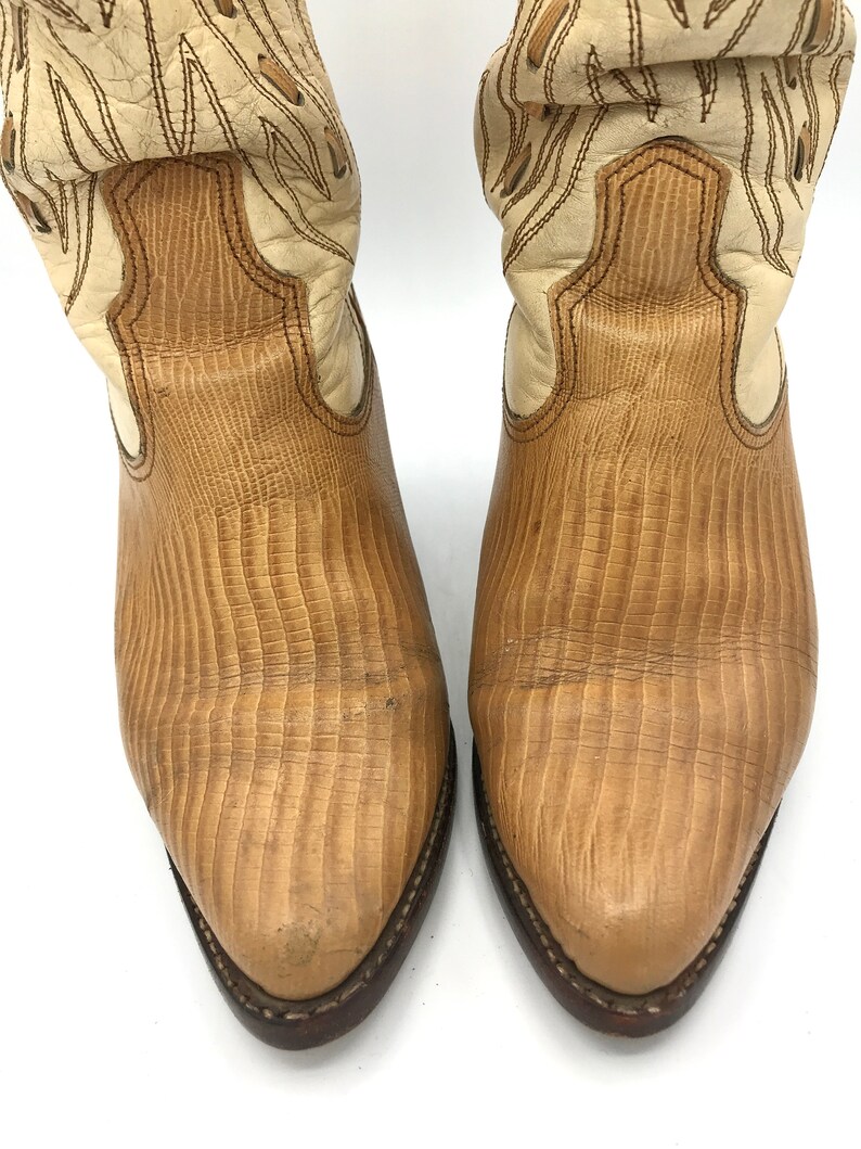 May include: A pair of cowboy boots with a light brown, textured leather design. The boots have a cream-colored upper with decorative stitching and a dark brown sole. The toe of the boot is pointed.
