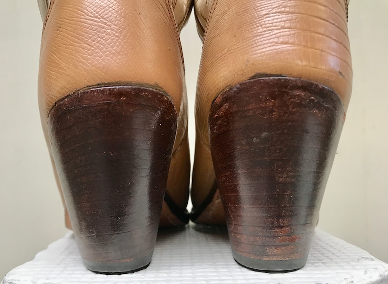 May include: Close-up of the heels of a pair of tan leather cowboy boots. The heels are dark brown, with a stacked design. The boots have a textured leather upper and are set against a neutral background. The boots appear to be well-worn.