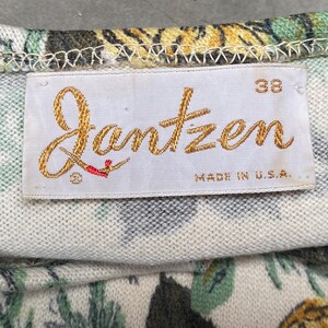 May include: A white fabric garment featuring a floral pattern in shades of green, yellow, and black. A rectangular white tag is stitched to the fabric, displaying the gold embroidered text Jantzen, the number 38, and the text MADE IN U.S.A.