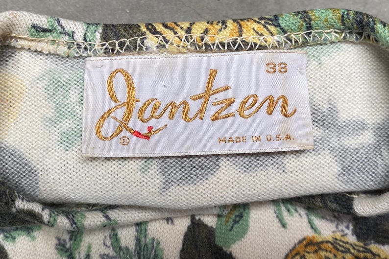 May include: A white fabric garment featuring a floral pattern in shades of green, yellow, and black. A rectangular white tag is stitched to the fabric, displaying the gold embroidered text Jantzen, the number 38, and the text MADE IN U.S.A.