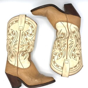 May include: A pair of beige and cream-colored cowboy boots. The boots feature decorative stitching, a pointed toe, and a stacked heel. The shaft is cream-colored with beige accents, and the foot is beige with a textured pattern. The heels are dark brown.