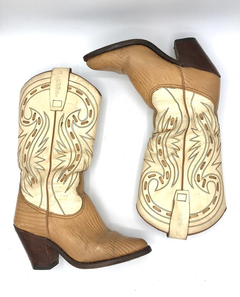 May include: A pair of beige and cream-colored cowboy boots. The boots feature decorative stitching, a pointed toe, and a stacked heel. The shaft is cream-colored with beige accents, and the foot is beige with a textured pattern. The heels are dark brown.