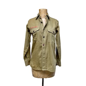 May include: Olive green button-down shirt with two chest pockets and a collar. The shirt has a red patch that reads "BOY SCOUTS OF AMERICA". The shirt is displayed on a vintage dress form.