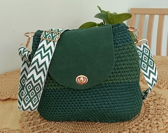 May include: A green crochet bag with a flap closure and a patterned strap. The bag has a two-tone design, with a darker green base and a lighter green top. The strap is white with a green zigzag pattern. The bag is handmade.