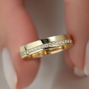 Puede incluir: Un anillo de banda de oro con una fila de pequeñas piedras preciosas claras engastadas a lo largo de los bordes superior e inferior. El anillo se sostiene entre los dedos, mostrando su superficie pulida y el brillo de las piedras. Un diseño clásico.