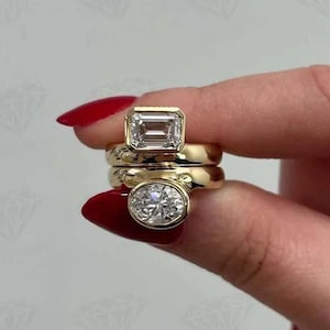 Puede incluir: Tres anillos de oro con piedras preciosas transparentes. El anillo superior presenta una piedra de talla esmeralda, el anillo central es una banda lisa y el anillo inferior tiene una piedra de talla ovalada. Los anillos están apilados.