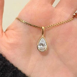 May include: A gold necklace with a pear-shaped pendant. The pendant features a large, clear gemstone with a halo of smaller stones. The chain is a delicate, box-style link. The necklace is displayed on a light skin tone.