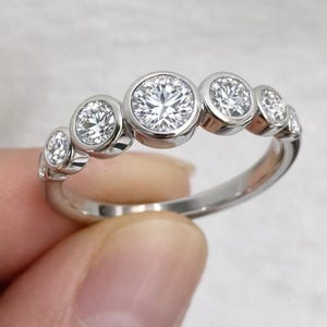 Op de afbeelding: Een zilveren ring met zes heldere, ronde edelstenen in individuele bezels. De edelstenen variëren in grootte, met de grootste in het midden. De ring heeft een gepolijste afwerking en staat tegen een neutrale achtergrond.