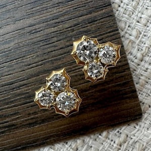 May include: A pair of gold stud earrings, each with three clear, round gemstones. The earrings have a distinctive, stylized design with a gold setting. The earrings are presented on a dark wooden surface.