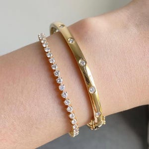 Könnte beinhalten: Zwei goldene Armbänder an einem Handgelenk. Eines ist ein Tennisarmband mit kleinen, runden, klaren Steinen. Das andere ist ein goldener Armreif mit mehreren größeren, klaren Steinen. Heller Hintergrund.