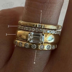 Puede incluir: Una colección de anillos de oro apilados en un dedo. Los anillos incluyen una banda con un solo diamante, una banda con pequeños diamantes, una banda gruesa con un diamante talla esmeralda y una banda con diamantes engastados.