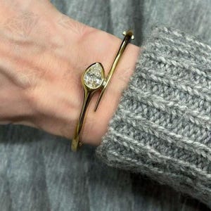 Op de afbeelding: Een goudkleurige armband met een peervormige diamant gezet in een bezel. De armband heeft een uniek, asymmetrisch ontwerp, met een gebogen band die om de pols wikkelt. De diamant schittert tegen het goud.