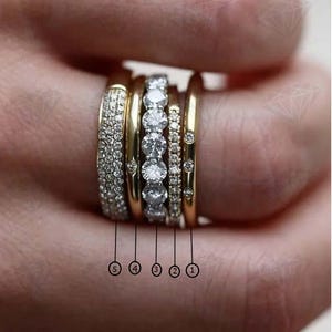 May include: A collection of five gold rings, each with a unique design. One ring is fully encrusted with small, clear stones, while another features a row of larger, clear stones. The other rings have a simple gold band design.