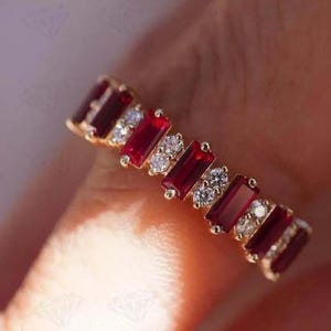 May include: A gold ring featuring alternating rectangular red gemstones and small, round, clear gemstones. The ring's design is set against a soft, blurred background, highlighting the jewelry's details.