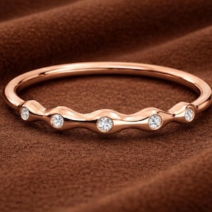 May include: A rose gold ring with five small, clear gemstones set in a wavy design. The ring is displayed on a brown, textured surface, showcasing its polished finish and elegant simplicity. The gemstones sparkle, adding a touch of luxury.