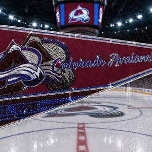 May include: A maroon pennant featuring the Colorado Avalanche logo and team name in sparkling embellishments. The pennant has a blue border and the text "EST. 1995". The background shows a hockey rink.