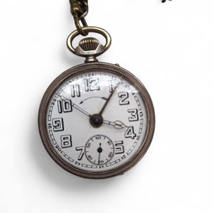 Op de afbeelding: Antieke zilveren zakhorloge met een witte wijzerplaat, zwarte cijfers en een kleine secondenwijzer. Het horloge heeft een ketting en een kroon. De wijzerplaat geeft de tijd weer.