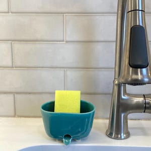 May include: A teal ceramic sponge holder with a small drainage spout at the front, featuring a bright yellow rectangular sponge placed inside. The holder has a smooth, glossy finish and is positioned next to a stainless steel kitchen faucet.