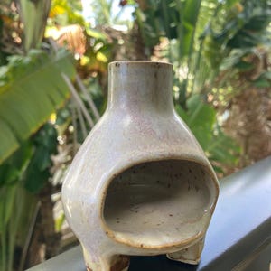 May include: A ceramic vessel with a wide, rounded body, a narrow neck, and a large, open arched cutout on the front. The piece features a speckled, light-colored glaze with subtle pink and lavender undertones, supported by three small feet.