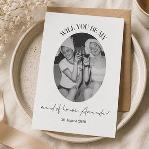 Könnte beinhalten: Eine Hochzeitseinladungskarte mit dem Text "WILL YOU BE MY maid of honor, Amanda? 26 August 2026" liegt auf einem Teller mit einer Tasse Kaffee und Schmuck. Die Karte zeigt ein Schwarz-Weiß-Foto von zwei Frauen.