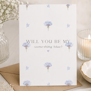 Op de afbeelding: Een witte wenskaart met een herhalend patroon van lichtblauwe korenbloemillustraties. De tekst in het midden luidt, WILL YOU BE MY something blue? met een klein lichtblauw hartsymbool onder de tekst op de kaart geplaatst.