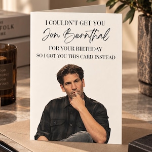 May include: A white greeting card featuring a photograph of actor Jon Bernthal wearing a dark button-down shirt. The text on the card reads: I COULDN'T GET YOU Jon Bernthal FOR YOUR BIRTHDAY SO I GOT YOU THIS CARD INSTEAD. The card is standing upright.