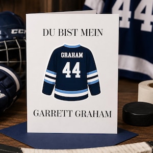 Könnte beinhalten: Eine weiße Grußkarte mit einer Illustration eines dunkelblauen Eishockeytrikots mit dem Namen GRAHAM und der Nummer 44 in Weiß. Der Text DU BIST MEIN steht über dem Trikot und der Name GARRETT GRAHAM ist in Schwarz unter dem Trikot aufgedruckt.
