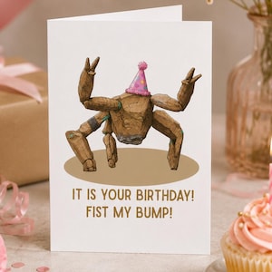 May include: A white greeting card featuring a brown, rock-like creature with multiple limbs wearing a pink polka-dot party hat. The text on the card reads, IT IS YOUR BIRTHDAY! FIST MY BUMP! The creature is positioned above the text on a tan oval.
