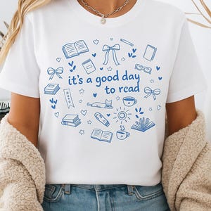 Könnte beinhalten: Weißes T-Shirt mit blauem Aufdruck "it's a good day to read". Das Design umfasst Bücher, Schleifen, Brillen und eine Katze. Das T-Shirt wird mit blauen Jeansshorts und einer beigen Jacke getragen.