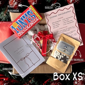 May include: A gift box labeled "Box XS" containing a chocolate bar, a candy cane, a card with "merry christmas" text, a recipe, a packet of cocoa with marshmallows, and a small gift tied with a red ribbon.
