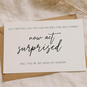 Könnte beinhalten: Eine weiße Karte mit dem Text "LET'S PRETEND LIKE YOU HAD NO IDEA THIS WAS COMING now act surprised WILL YOU BE MY MAID OF HONOR?" in schwarzer Schrift, auf einem braunen Umschlag. Eine beige Kerze und getrocknete Pflanzen sind im Hintergrund.