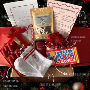 Könnte beinhalten: Eine weihnachtliche Geschenkbox mit verschiedenen Artikeln. Enthält eine Karte, heiße Schokolade mit Marshmallows, eine Kerze, Socken mit Weihnachtsmotiv, eine Schokoladentafel, ein rotes Scrunchie und eine Zuckerstange.