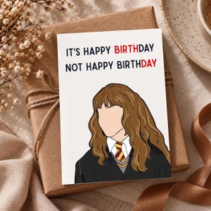 May include: A white greeting card featuring a cartoon illustration of a person with long wavy brown hair wearing a black robe, white collared shirt, and a red and yellow striped tie. The text reads: IT'S HAPPY BIRTHDAY NOT HAPPY BIRTHDAY.