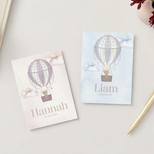 Op de afbeelding: Twee geboortekaartjes met illustraties van luchtballonnen. Eén kaart heeft een roze achtergrond met de naam "Hannah" en de datum. De andere kaart heeft een blauwe achtergrond met de naam "Liam" en de datum. Een gouden pen ligt op tafel.