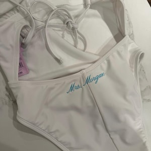 May include: A white one-piece swimsuit featuring thin spaghetti straps that tie at the back. The garment is embroidered with the text Mrs. Morgan in a light blue cursive script. A small purple product tag is attached to the side of the swimsuit.