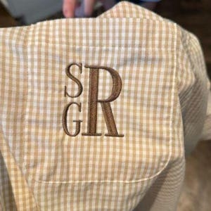 Puede incluir: Primer plano de un bolsillo de camisa a cuadros beige y blanco con las iniciales bordadas "SGR" en hilo marrón. La camisa tiene un patrón de cuadros clásico. El bolsillo está cosido cuidadosamente a la tela, con una estética personalizada.