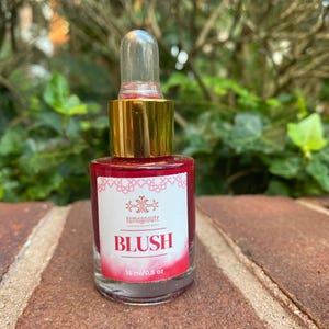 Puede incluir: Una pequeña botella de rubor de vidrio con una tapa dorada y un cuentagotas transparente. La etiqueta dice "BLUSH" en letras grandes y en negrita. La botella contiene 15 ml de líquido rosa. El fondo está borroso con follaje verde.