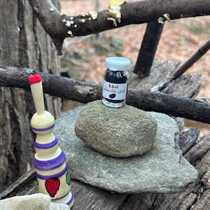 May include: A small glass jar with a white lid, filled with black powder, sits on a gray stone. The jar has a label with text in a foreign language. Beside it is a decorative wooden object with purple and red accents.