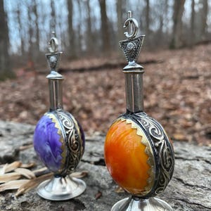 Puede incluir: Dos frascos de perfume decorativos con detalles metálicos plateados. Uno tiene un diseño veteado morado y blanco, el otro es naranja y amarillo. Ambos tienen adornos de metal y una parte superior decorativa. Los frascos se exhiben al aire libre.
