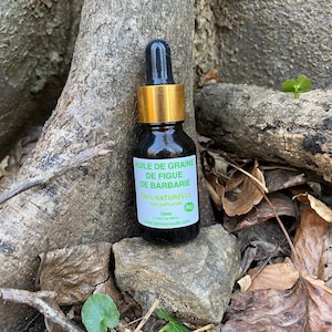 May include: A small, dark glass bottle with a black dropper and gold-colored cap, labeled in French. The label reads "HUILE DE GRAINE DE FIGUE DE BARBARIE 100% NATURELLE" and includes a bio certification. The bottle is set on a rock.