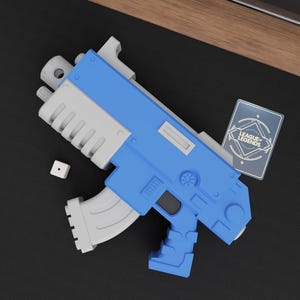 May include: A blue and gray toy gun with a die and a League of Legends card. The toy gun has a blue body and gray accents. The League of Legends card is rectangular with the game's logo.