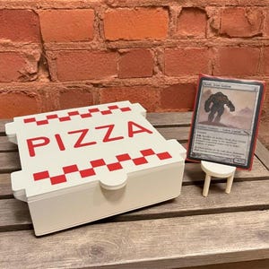 Puede incluir: Una caja de pizza blanca con texto rojo "PIZZA" y detalles a cuadros rojos. Una tarjeta coleccionable se muestra en un pequeño soporte blanco. El fondo es una pared de ladrillos y una mesa de madera.