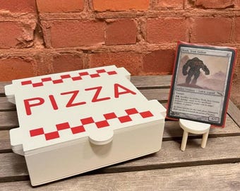 Op de afbeelding: Een witte pizzadoos met rode tekst "PIZZA" en rode geblokte accenten. Een ruilkaart wordt op een kleine witte standaard gepresenteerd. De achtergrond is een bakstenen muur en een houten tafel.